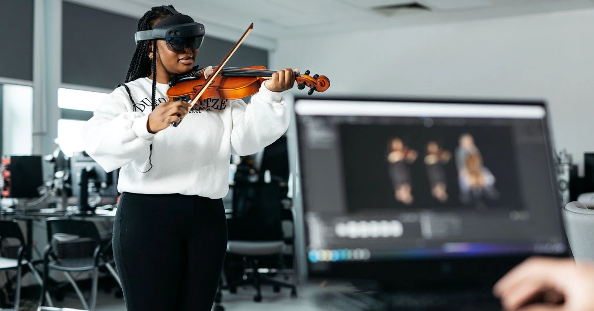 A violinist wearing an augmented reality headset beside a screen showing virtual ensemble musicians